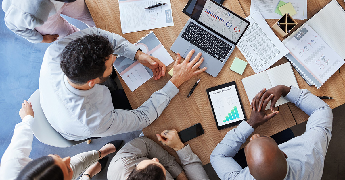 A group of business leaders gather around a table with paper and computers